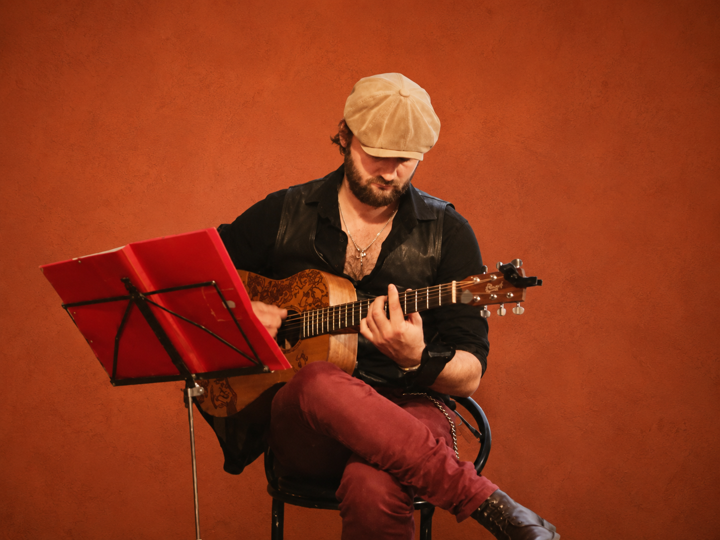 Jean-Michel Burckle assis avec sa guitare acoustique devant un pupitre rouge, fond terracotta